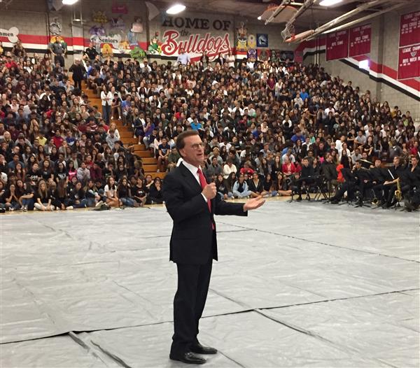 Lowel Milken, an adult male, stands in the center of a high school gym as he speaks. Students watch from bleachers.